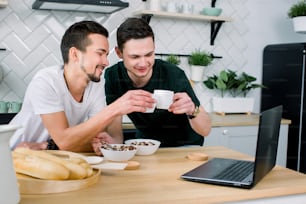 Two young handsome men having breakfast and drinking coffee while using laptop in the kitchen at the morning. Smiling men watching film using laptop and having breakfast