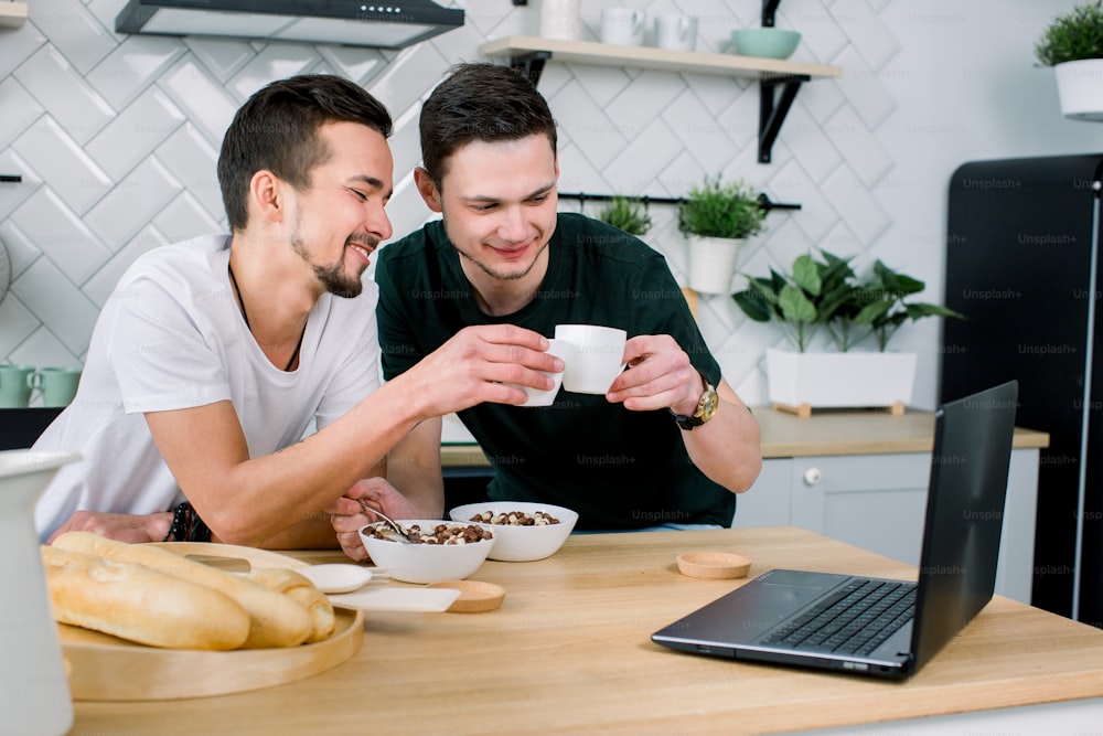 Dois jovens bonitos tomando café da manhã e tomando café enquanto usavam laptop na cozinha pela manhã. Homens sorridentes assistindo filme usando laptop e tomando café da manhã