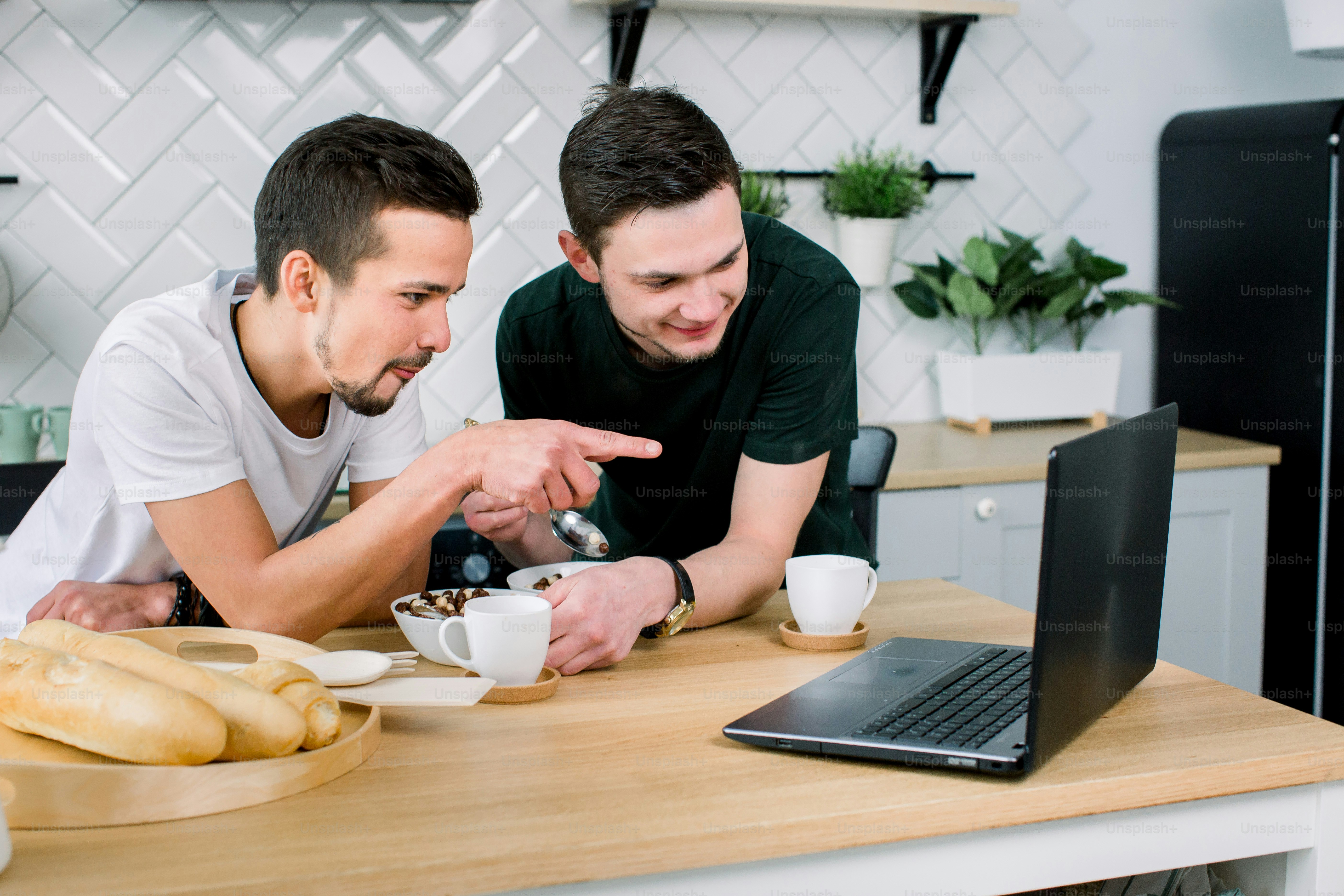 Two young handsome men having breakfast and drinking coffee while using laptop in the kitchen at the morning. Smiling men watching film using laptop and having breakfast