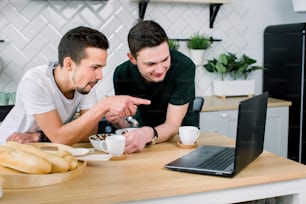 아침에 부엌에서 노트북을 사용하면서 아침을 먹고 커피를 마시는 두 젊은 잘 생긴 남자. 노트북을 사용하여 영화를 보고 아침을 먹는 웃는 남자들