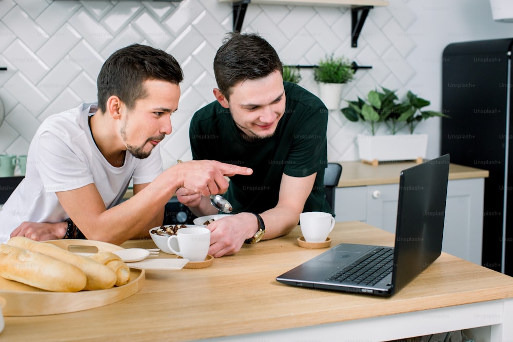 Dos hombres jóvenes y guapos desayunando y bebiendo café mientras usan una computadora portátil en la cocina por la mañana. Hombres sonrientes viendo películas usando una computadora portátil y desayunando