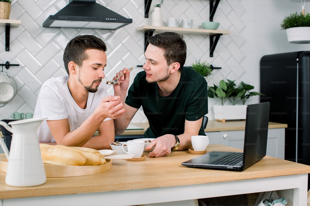 Male gay couple sitting at the table and having breakfast in their kitchen. Handsome young man in black t-shirt feeding his partner. Gay couple