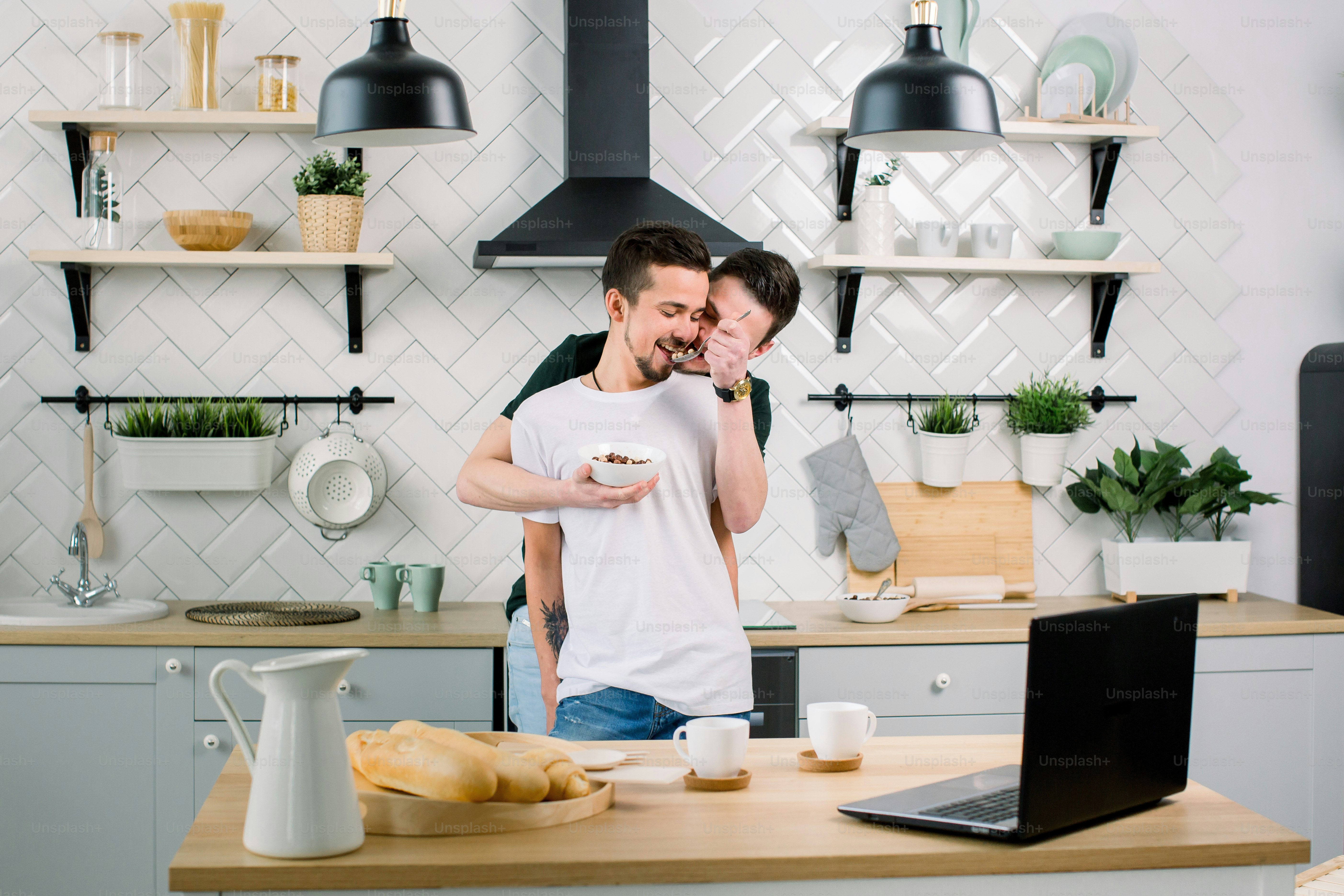 Gay couple hugging in the kitchen, standing at the dinner table, one man feeding another and having fun. People, homosexuality, same-sex family, gay relationships concept.