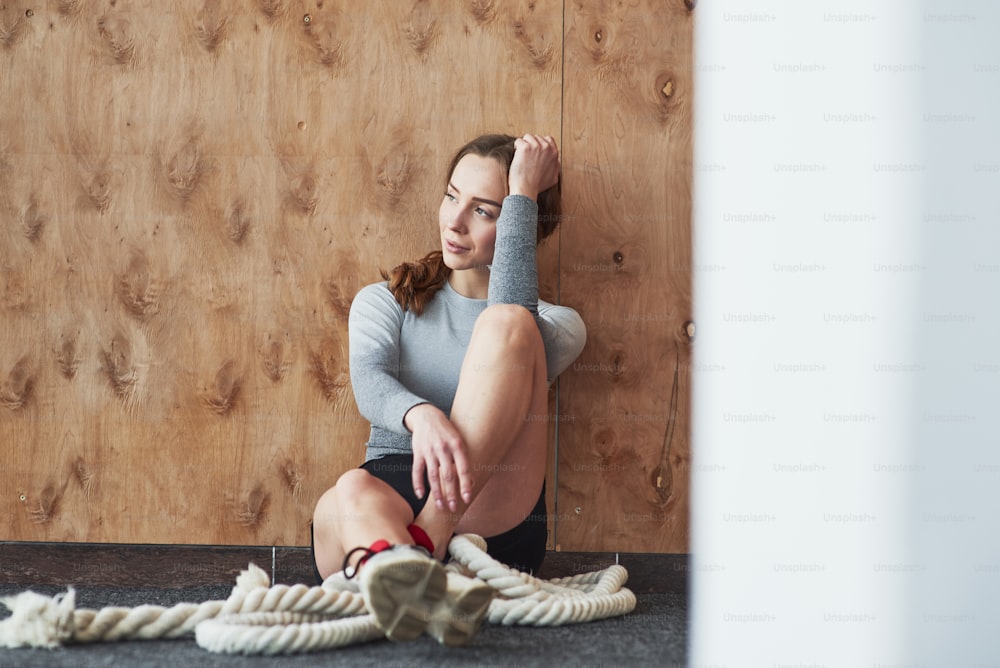 Leaning on the wooden wall. Sportive young woman have fitness day in the gym at morning time.