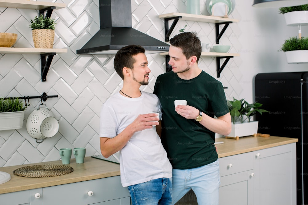 Pareja gay feliz con café en la cocina casera, primer plano. Hombres homosexuales guapos bebiendo café y mirándose en la cocina