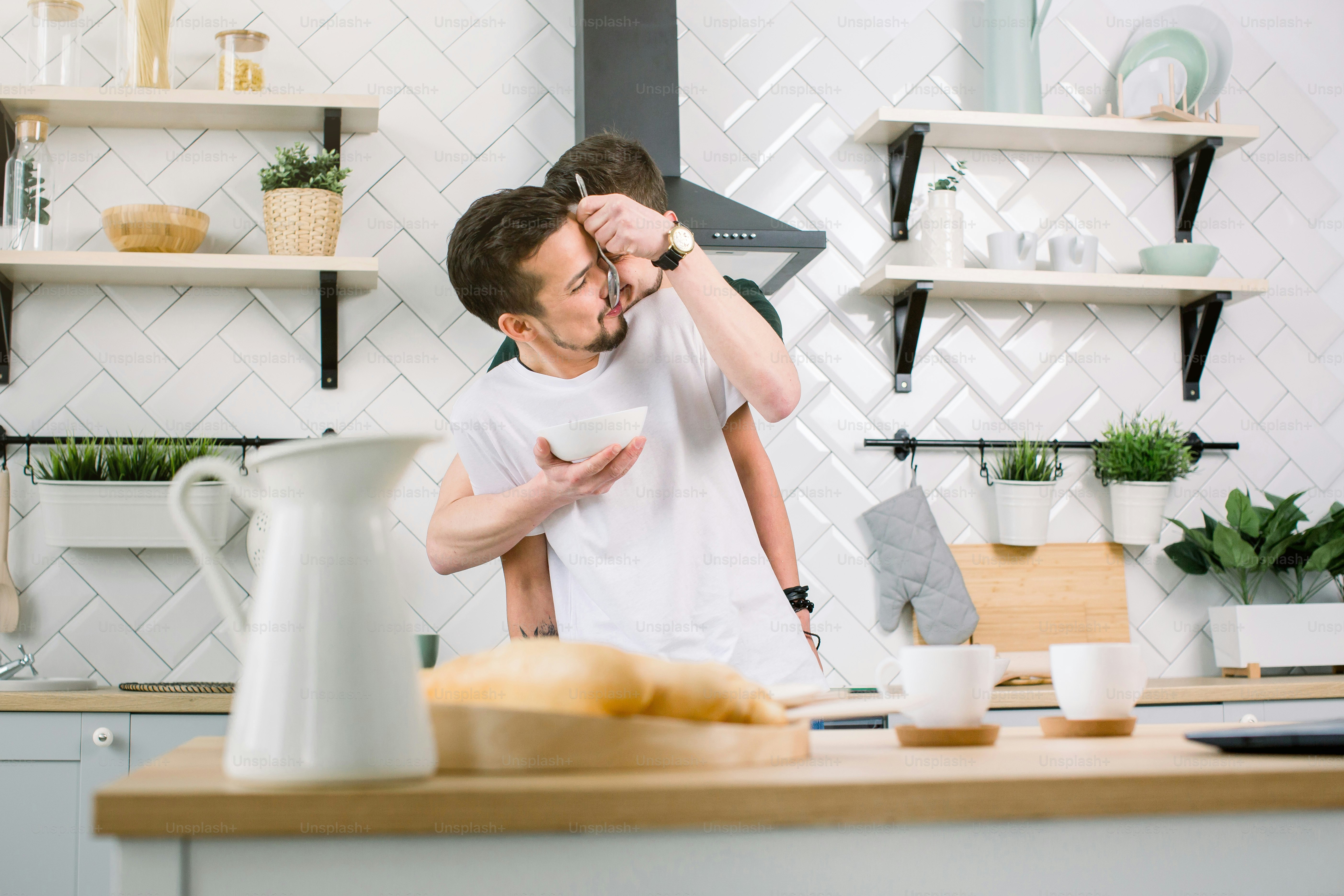 Gay couple hugging in the kitchen, standing at the dinner table, one man feeding another and having fun. People, homosexuality, same-sex family, gay relationships concept.