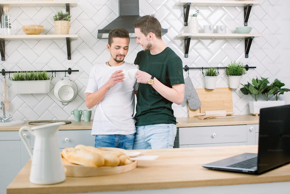 Casal gay feliz com café na cozinha de casa, close-up. Homens homossexuais bonitos tomando café e olhando uns para os outros na cozinha