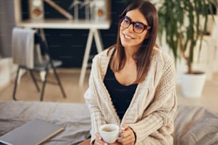 Attractive caucasian brunette in beige sweater and with eyeglasses sitting on the bed in bedroom in morning.