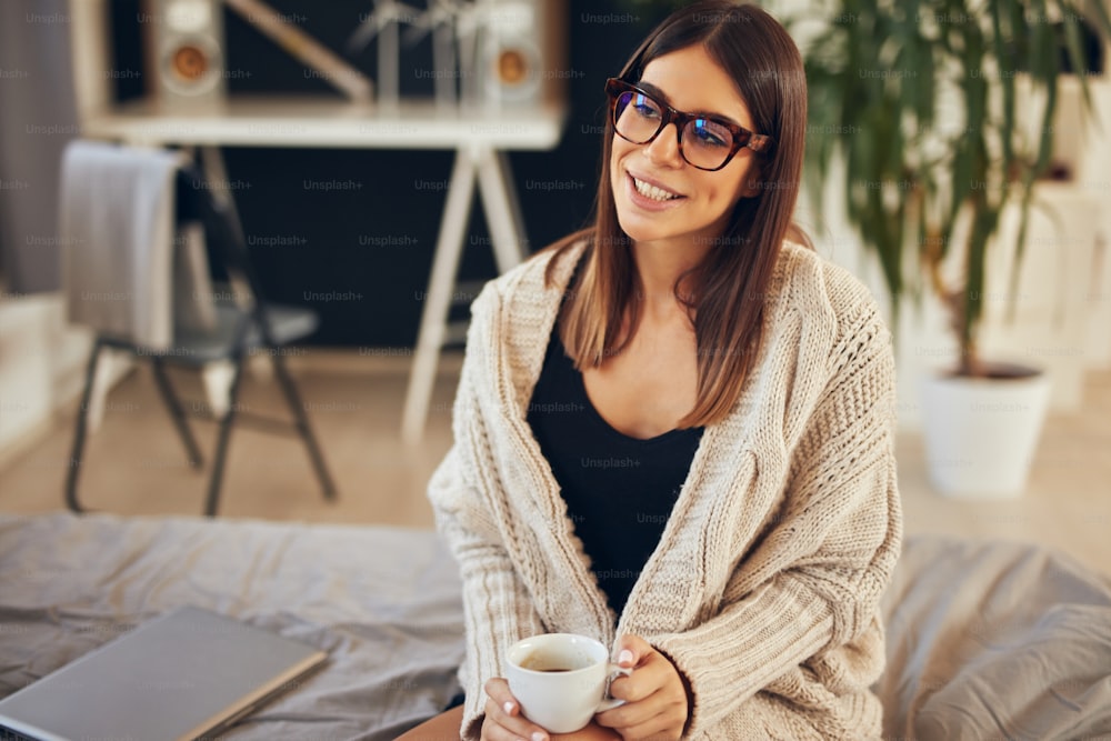 Attraente bruna caucasica in maglione beige e con occhiali da vista seduta sul letto in camera da letto al mattino.