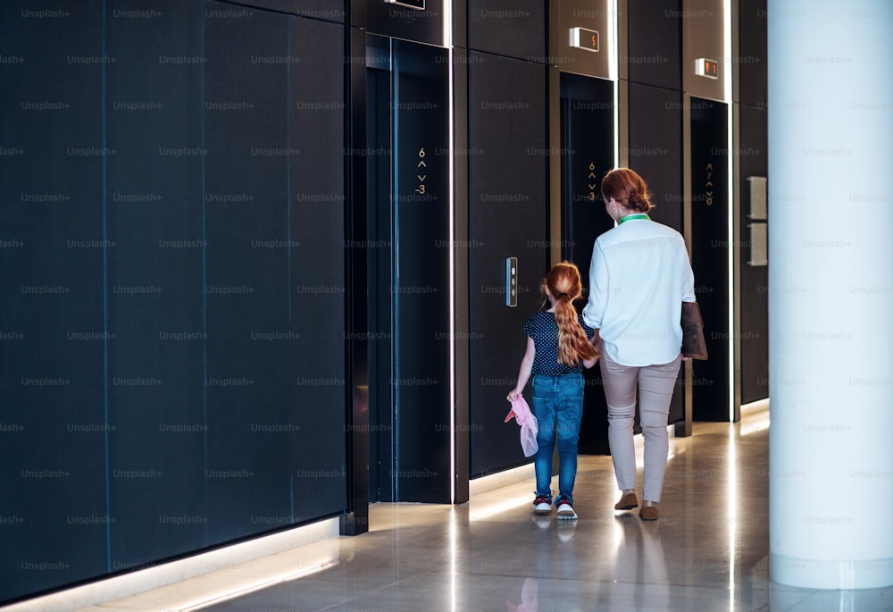 A side view of businesswoman with small daughter walking by elevators in office building.