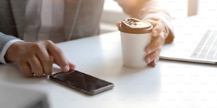 Ausschnittaufnahme eines professionellen Geschäftsmannes, der an seinem Projekt arbeitet, während er sein Smartphone berührt und eine Kaffeetasse hält