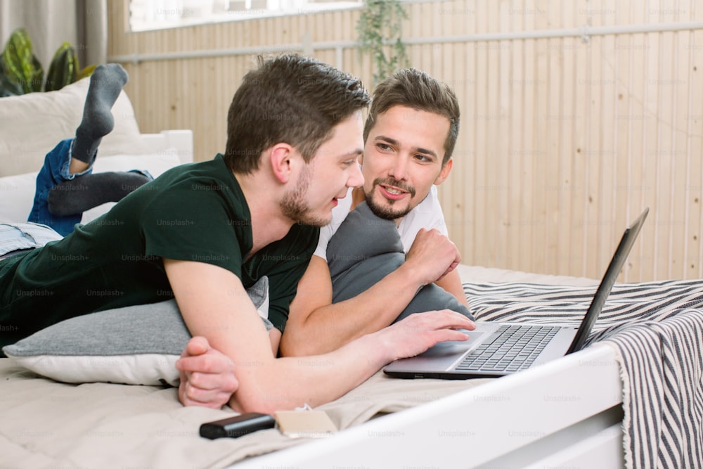 Smiling Caucasian gay couple using laptop on bed at home. Homosexual, gay couple concept