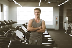 Young man in sportswear smiling and standing near the treadmill before or after training on gym. Cardio workout, running on treadmill. Healthy lifestyle, guy training in gym. Flare effect, copy space