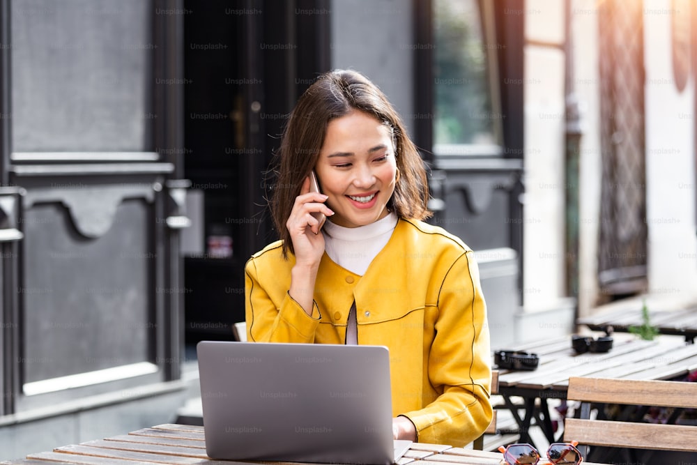 Happy entrepreneur working with a phone and laptop in a coffee shop in the street. Beautiful young Asian girl working at a coffee shop with a laptop.