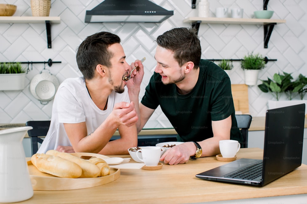 Male gay couple sitting at the table and having breakfast in their kitchen. Handsome young man in black t-shirt feeding his partner. Gay couple