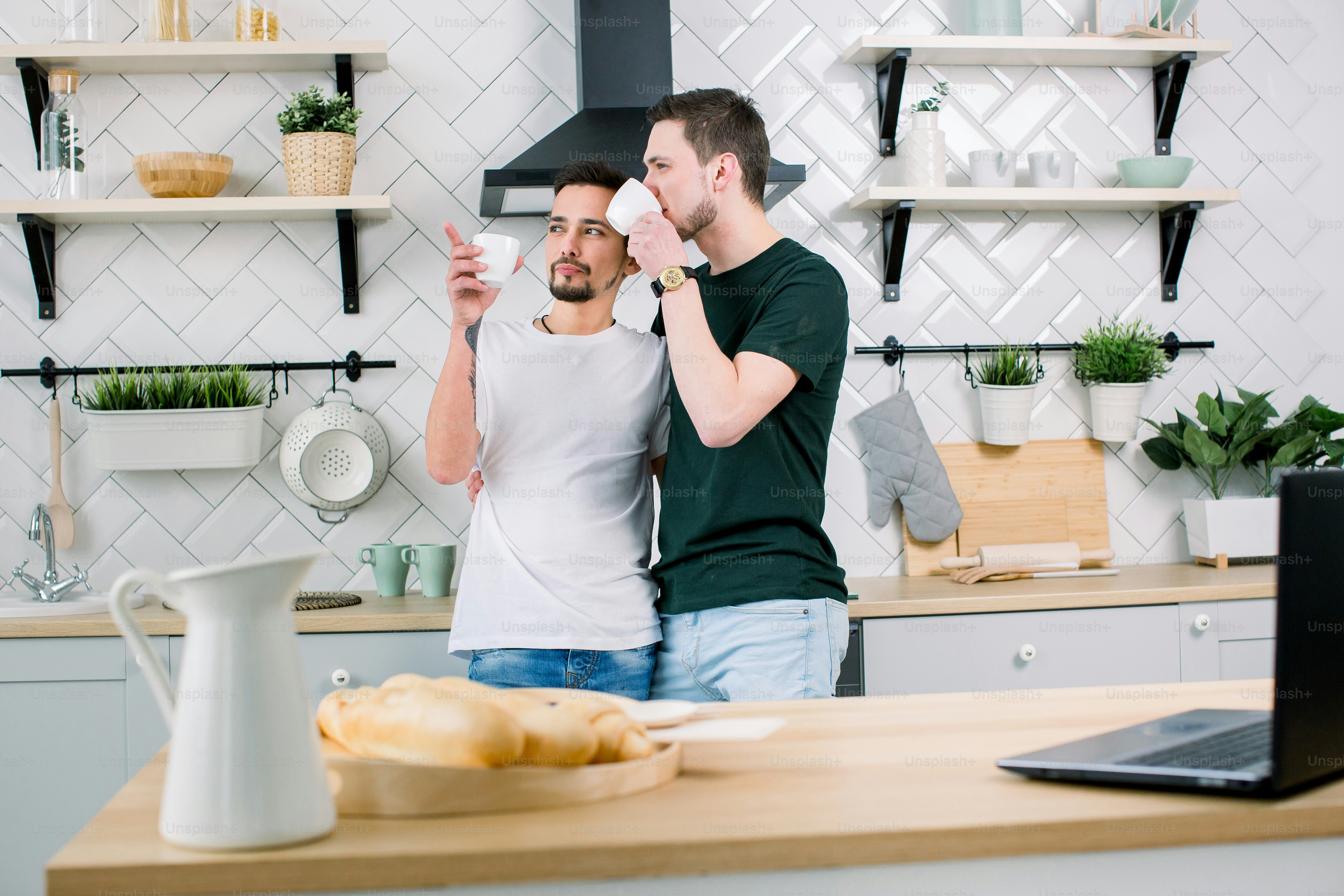 People, homosexuality, same-sex family, gay relationships concept. Male gay couple hugging each other and drinking their morning coffee while standing at the kitchen at home