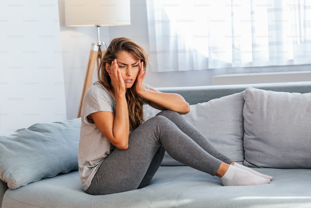 Porträt einer attraktiven Frau, die zu Hause auf einem Sofa sitzt, mit Kopfschmerzen, Schmerzen und einem Ausdruck von Unwohlsein. Verärgerte depressive Frau, die auf der Couch liegt und starke Kopfschmerzen Migräne verspürt.
