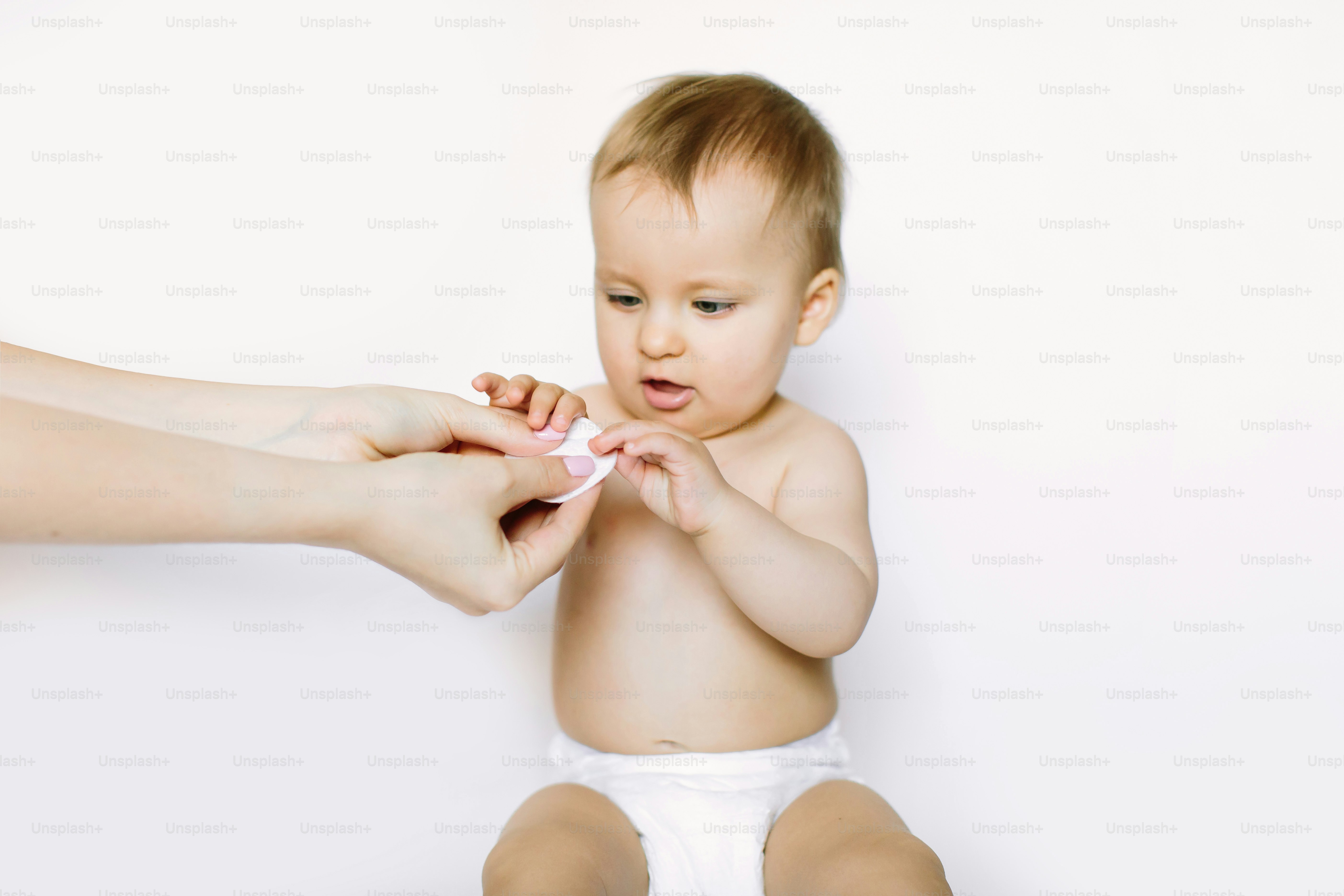 Wadded cotton pad in mother hand on a white background isolation. Cute little baby girl sitting and looking at the mother hands and cotton pad. Baby hygiene concept.