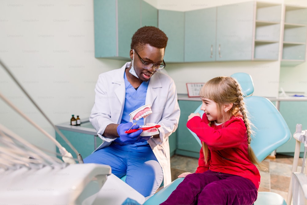 행복한 미소 짓는 흑인 치과 의사는 백인 어린 소녀에게 양치질하는 방법을 알려줍니다. 충치 예방, 치과, 치아 위생 개념
