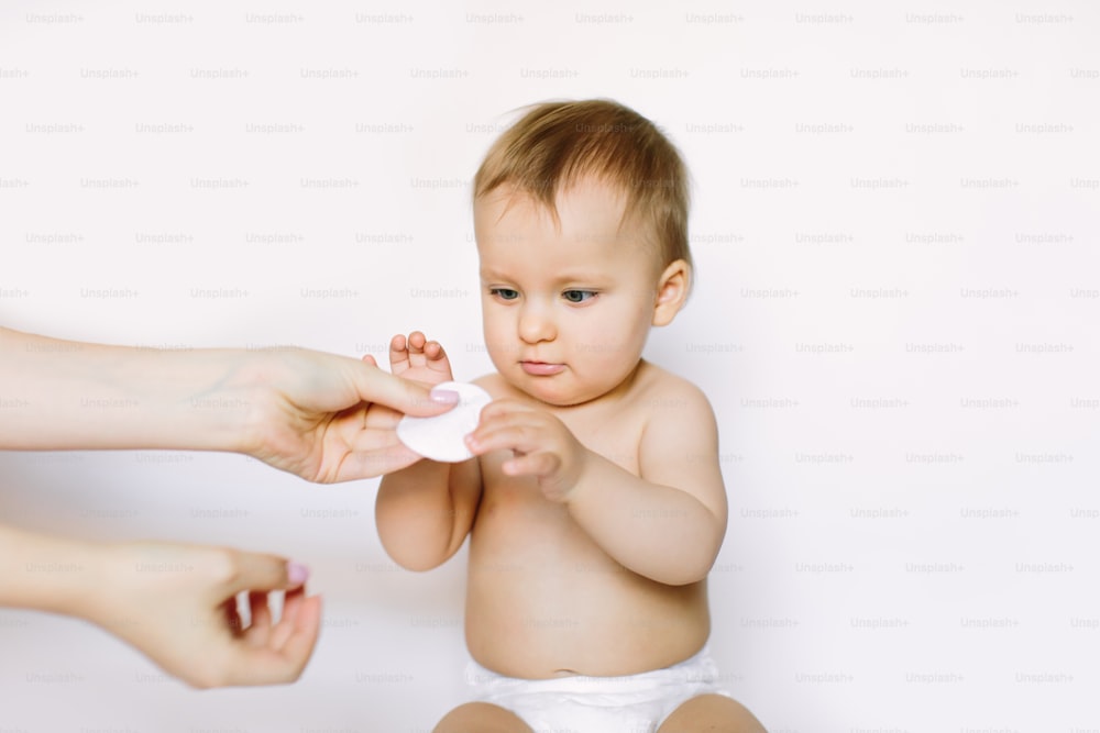 Tampon de coton ouaté dans la main mère sur un fond blanc isolant. Mignonne petite fille assise et regardant les mains de la mère et le coton. Concept d’hygiène pour bébé.