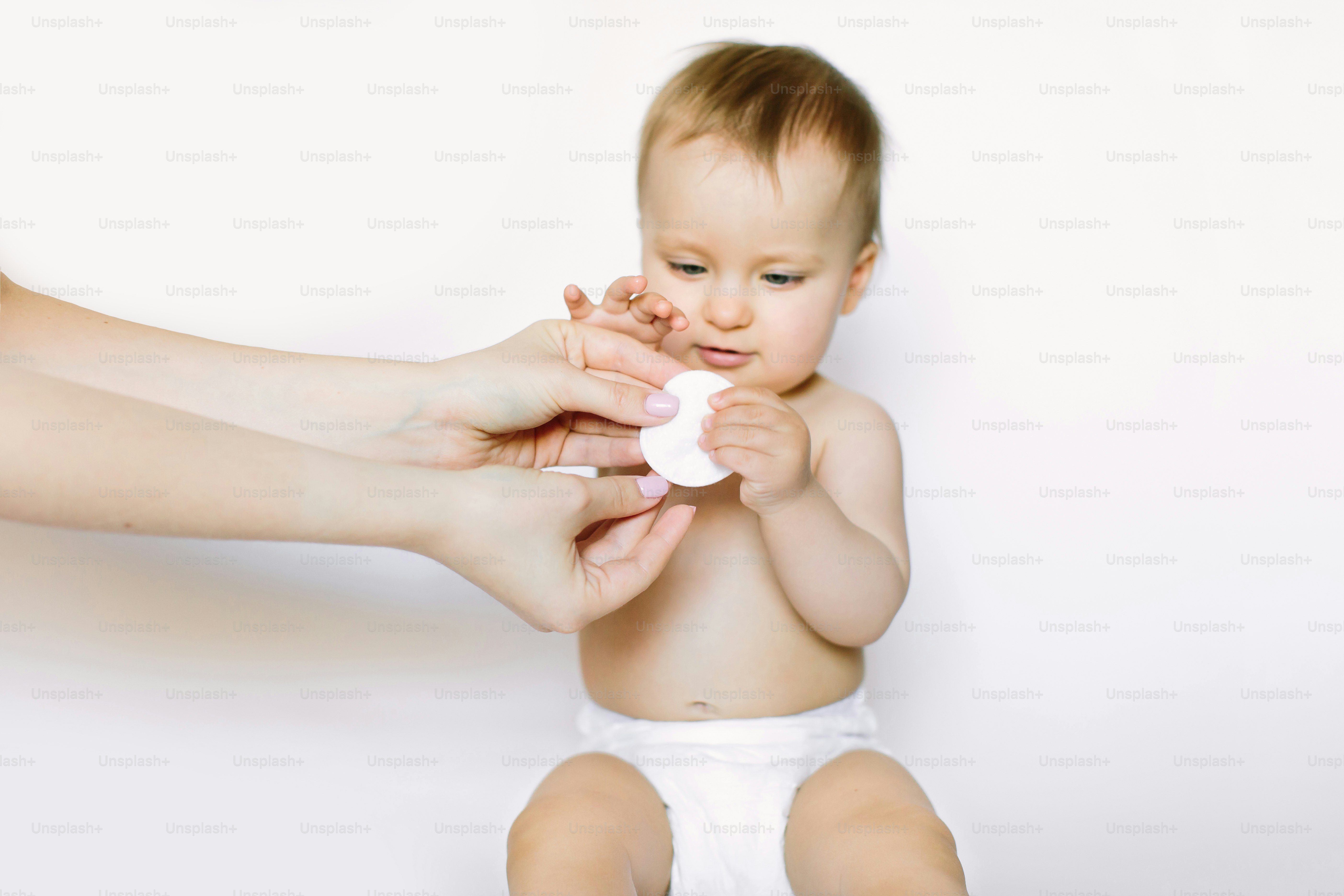Little happy baby girl of one year old in white diaper sitting and holding the white cotton pad together with her mother, isolated on white background. Baby car and hygiene concept