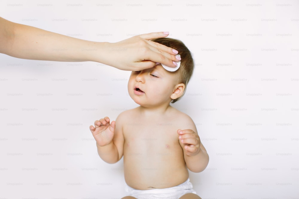 La main de la mère nettoie le visage d’une petite fille d’un an avec un coton sur fond blanc. Hygiène du bébé, soins, soins de santé, concept médical