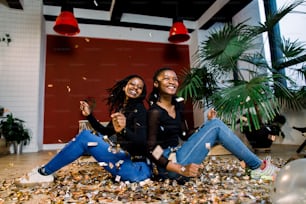 Deux filles africaines, des amis heureux et élégants célébrant le nouvel an ou une fête d’anniversaire s’assoient l’un avec l’autre et lancent des confettis. Les femmes d’élégance de la mode profitent du temps ensemble.