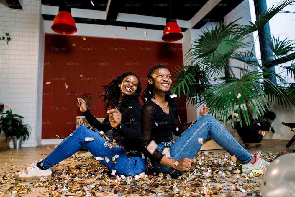 Two african girls, happy stylish friends celebrating new year or birthday party sit back with each other and throw a confetti. Fashion elegance women enjoying time together.