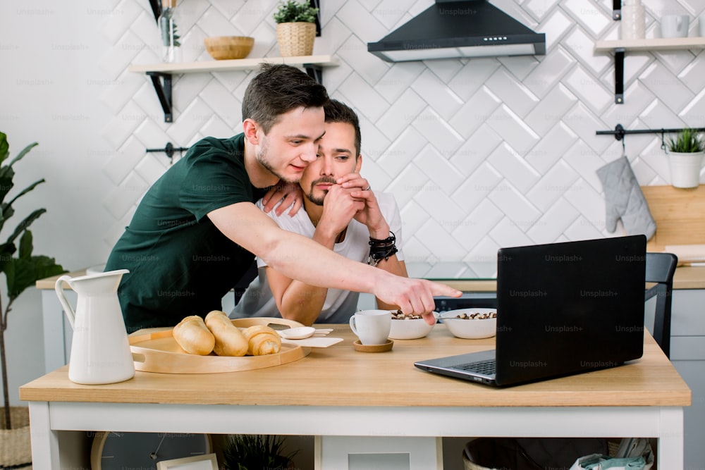 Attractive young Caucasian gay couple, homosexual couple, in kitchen, embracing and having breakfast at the dinner table, using internet on laptop. Studio, light kitchen background.