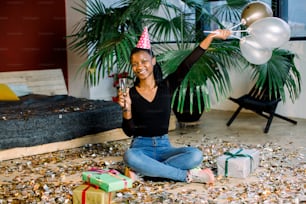 Fête d’anniversaire, carnaval du nouvel an. Jeune femme souriante africaine célébrant un événement lumineux, . Des confettis scintillants, s’amuser, danser et boire du champagne