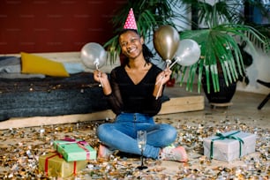 Funny African American girl in birthday hat and with air balloons sitting on the floor with present boxes gifts and confetti. Celebration, party concept