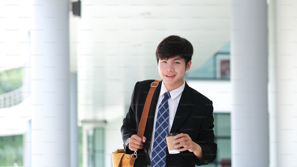 Handsome smiling young businessman wearing suit walking outdoor holding coffee cup while coffee break.