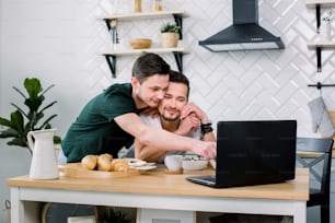 Attractive young Caucasian gay couple, homosexual couple, in kitchen, embracing and having breakfast at the dinner table, using internet on laptop. Studio, light kitchen background.