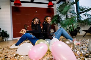 Dos chicas africanas, amigas felices y elegantes que celebran el año nuevo o la fiesta de cumpleaños se sientan juntas y lanzan un confeti. Moda elegancia mujeres disfrutando del tiempo juntas.