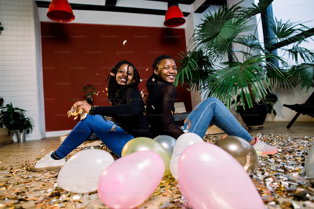 Two african girls, happy stylish friends celebrating new year or birthday party sit back with each other and throw a confetti. Fashion elegance women enjoying time together.