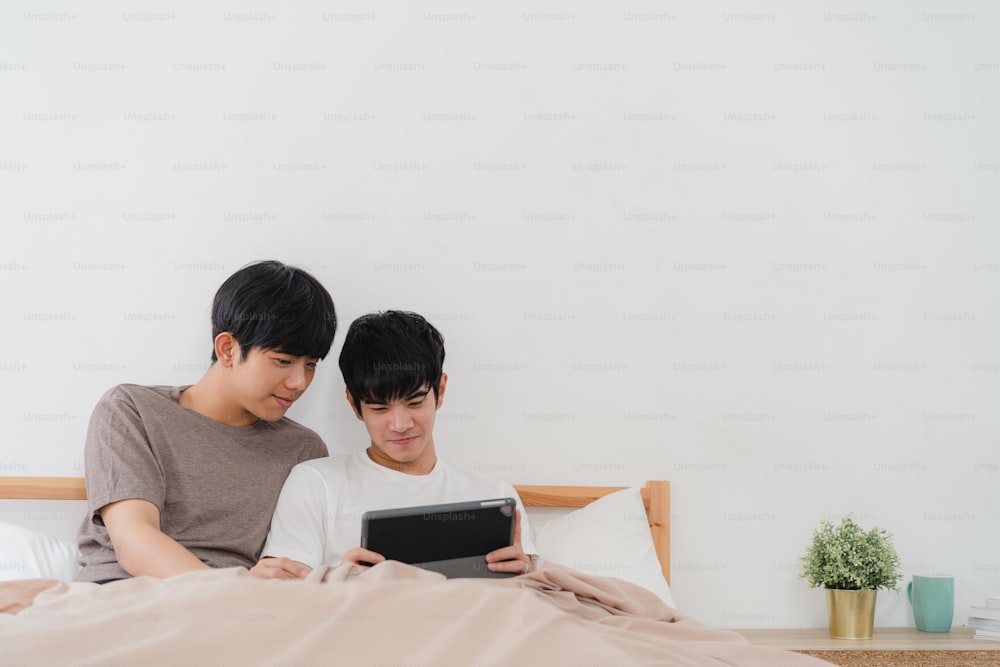 Asian Gay couple using tablet at home. Young Asian LGBTQ men happy relax rest together after wake up, check mail and social media lying on bed in bedroom at home in the morning concept.