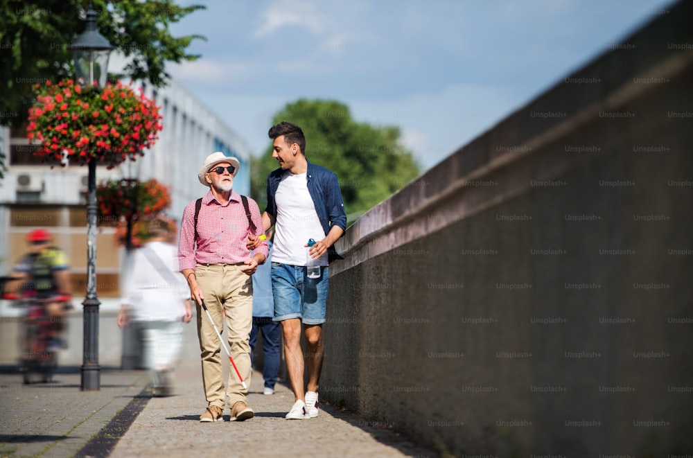A young man and blind senior with white cane walking on pavement in city, talking.