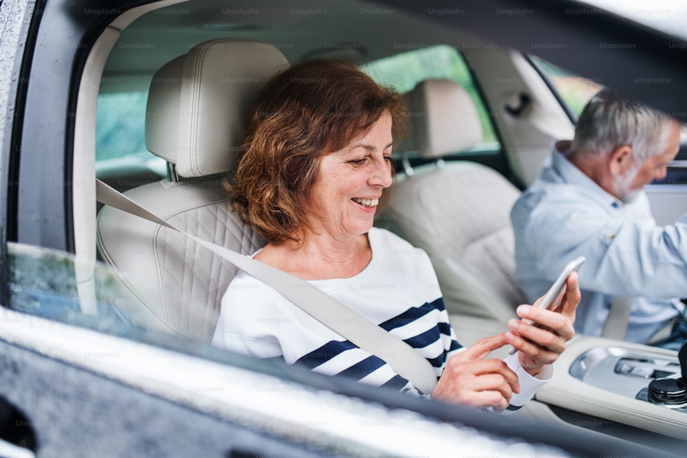 A happy senior couple with smartphone sitting in car, going on trip.