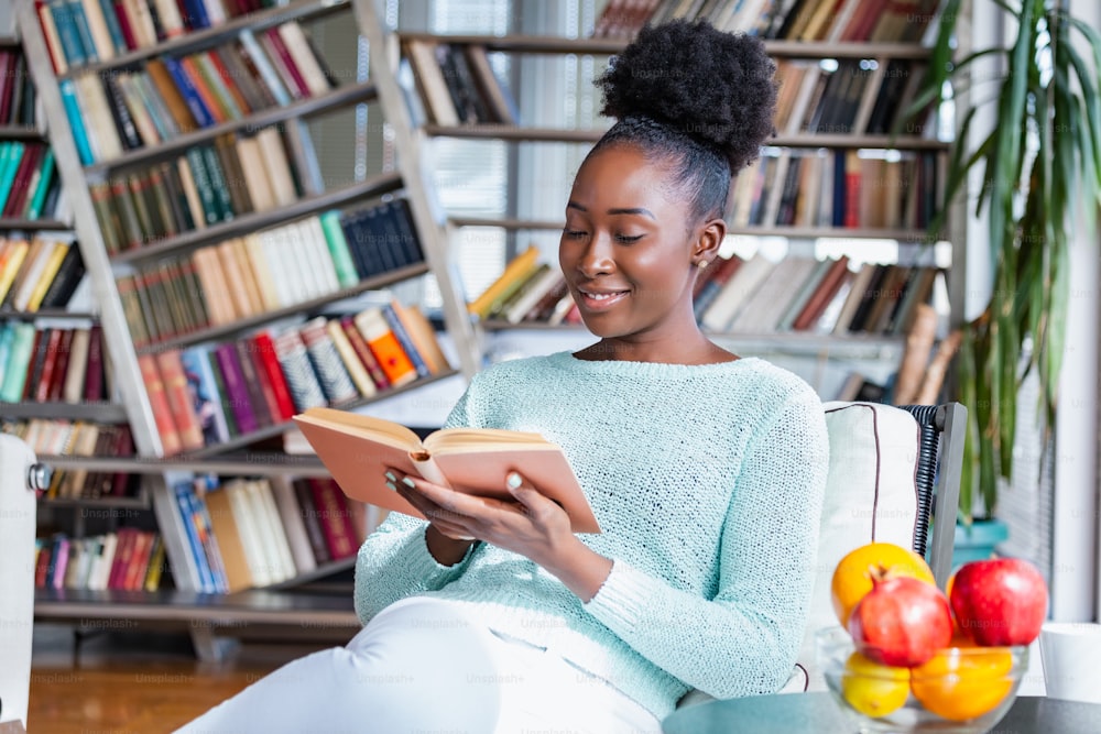 Joven hermosa chica afroamericana leyendo un libro en el sofá con las estanterías de la biblioteca en la parte de atrás. Mujer hermosa en un sofá blanco leyendo un libro