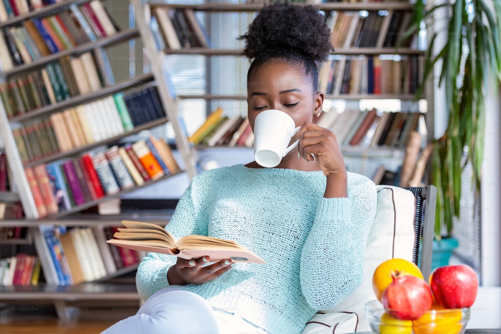 Jeune femme avec un livre et une tasse de boisson chaude. Jolie fille afro-américaine avec la lecture d’un livre assis dans une bibliothèque à la maison avec des étagères à l’arrière.