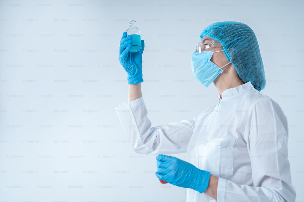 Low angle view of chemist woman in coat and protective gloves making laboratory analysis, holding medical glass bottle with blue liquid, standing isolated on white background with copy space