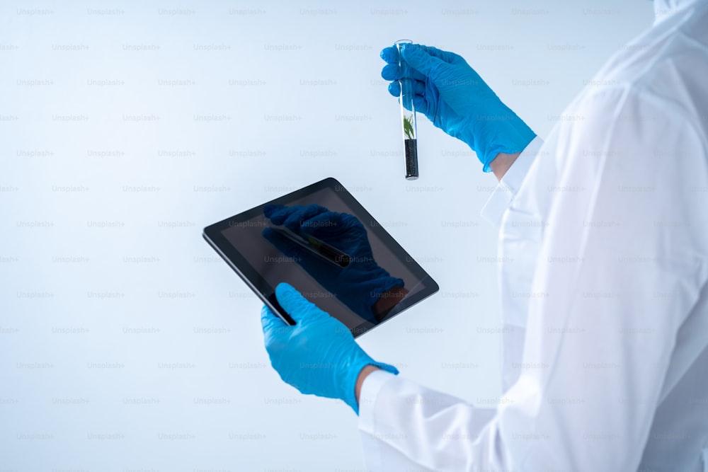 Cropped view of scientist woman in coat making laboratory research on tablet computer, holding test tube with green plant in soil, standing isolated on white background  with copy space