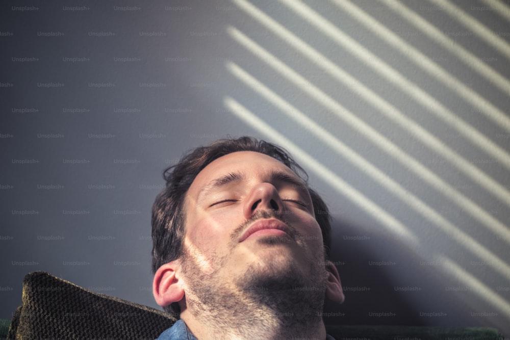 Portrait d’un homme caucasien somnolant sur le canapé, sur le visage tombe l’ombre des stores