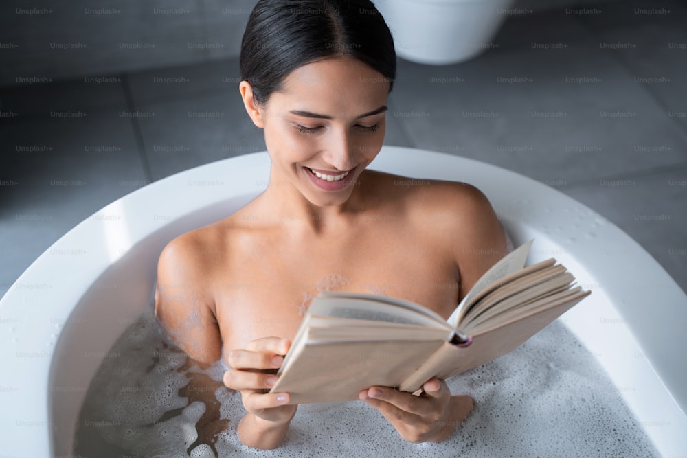 Vista superior de una mujer joven y bonita sonriente leyendo un libro en el baño