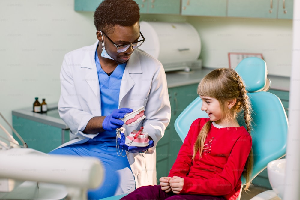 Little patient at the dentist's office. Young African American dentist talking with patient small girl holding artificial jaws in his hands.