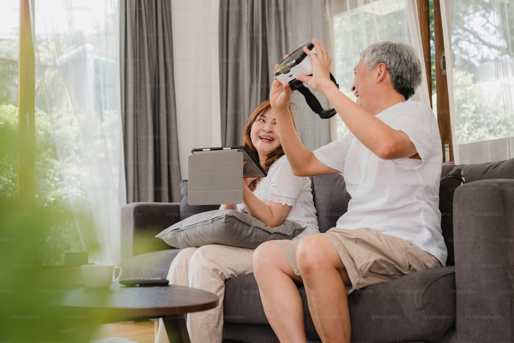 Asian elderly couple using tablet and virtual reality simulator playing games in living room, couple feeling happy using time together lying on sofa at home. Lifestyle Senior family at home concept.