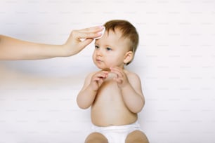 La mano de la madre limpia la cara de una niña de un año con una almohadilla de algodón sobre fondo blanco. Higiene, cuidado, atención médica, concepto médico del bebé