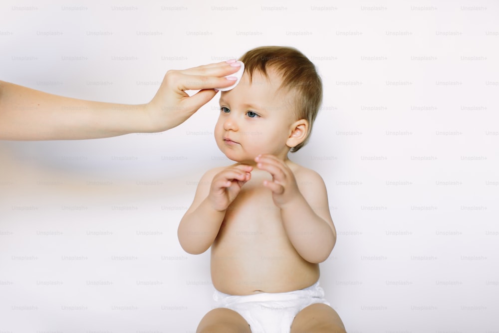 La main de la mère nettoie le visage d’une petite fille d’un an avec un coton sur fond blanc. Hygiène du bébé, soins, soins de santé, concept médical