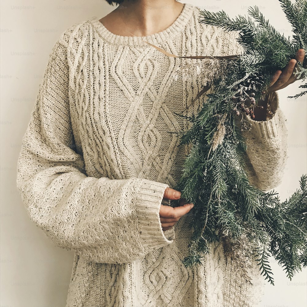 Girl in knitted sweater holding stylish rustic wreath. Hipster girl with rural modern wreath on white wall background. Festive decoration. Phone photo