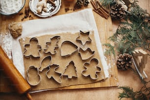 Making christmas gingerbread cookies, flat lay. Raw dough with metal cutters for cookies and wooden rolling pin, anise, ginger, cinnamon, pine cones, fir branches on rustic table.